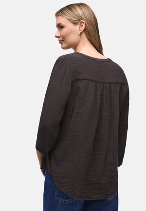 Femme aux cheveux blonds portant un chemisier noir texturé à manches longues et un jean bleu, de profil sur un fond clair uni.