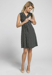 Black dress with white polka dots, sleeveless design, v-neckline, gathered waist, and a flared skirt. Model adjusts dress detail.