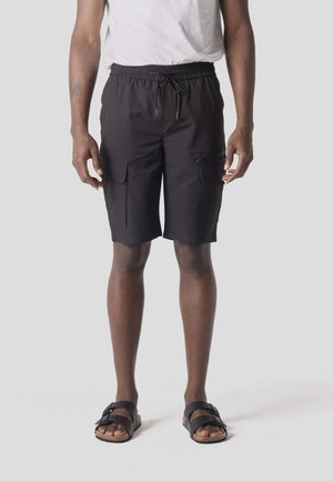 Férfi fekete oldalzsebes vászonnadrágban, fehér pólóban és fekete csatos szandálban, egyszínű háttér előtt állva.