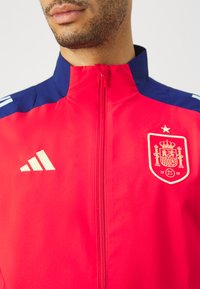 Chaqueta de entrenamiento roja con detalles en azul marino, que cuenta con cierre de cremallera, tres rayas blancas en el hombro y un escudo dorado de España.