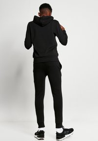 Sudadera negra con capucha y joggers a juego, fabricados en un tejido suave, con un corte regular, puños acanalados y bolsillos laterales. Llevado con zapatillas negras.