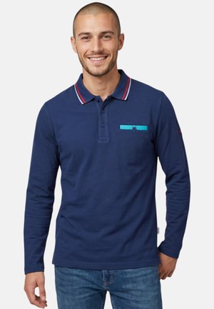 Homme portant un polo bleu marine à manches longues avec une bordure rouge et blanche au col et une poche poitrine bleue boutonnée, souriant.