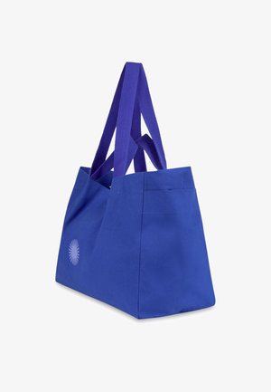 Blaue Leinentragetasche mit langen, lila Stoffgriffen, die auf einer Seite ein einfaches Sonnenstrahlen-Design zeigt. Robuste rechteckige Form mit zusätzlichen Seitentaschen.