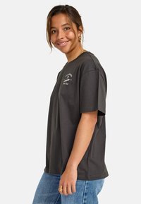 Ung kvinne som smiler, iført en mørk, oversized t-skjorte med et lite hvitt grafisk design, blå jeans, gulløredobber og ringer, mot en enkel bakgrunn.