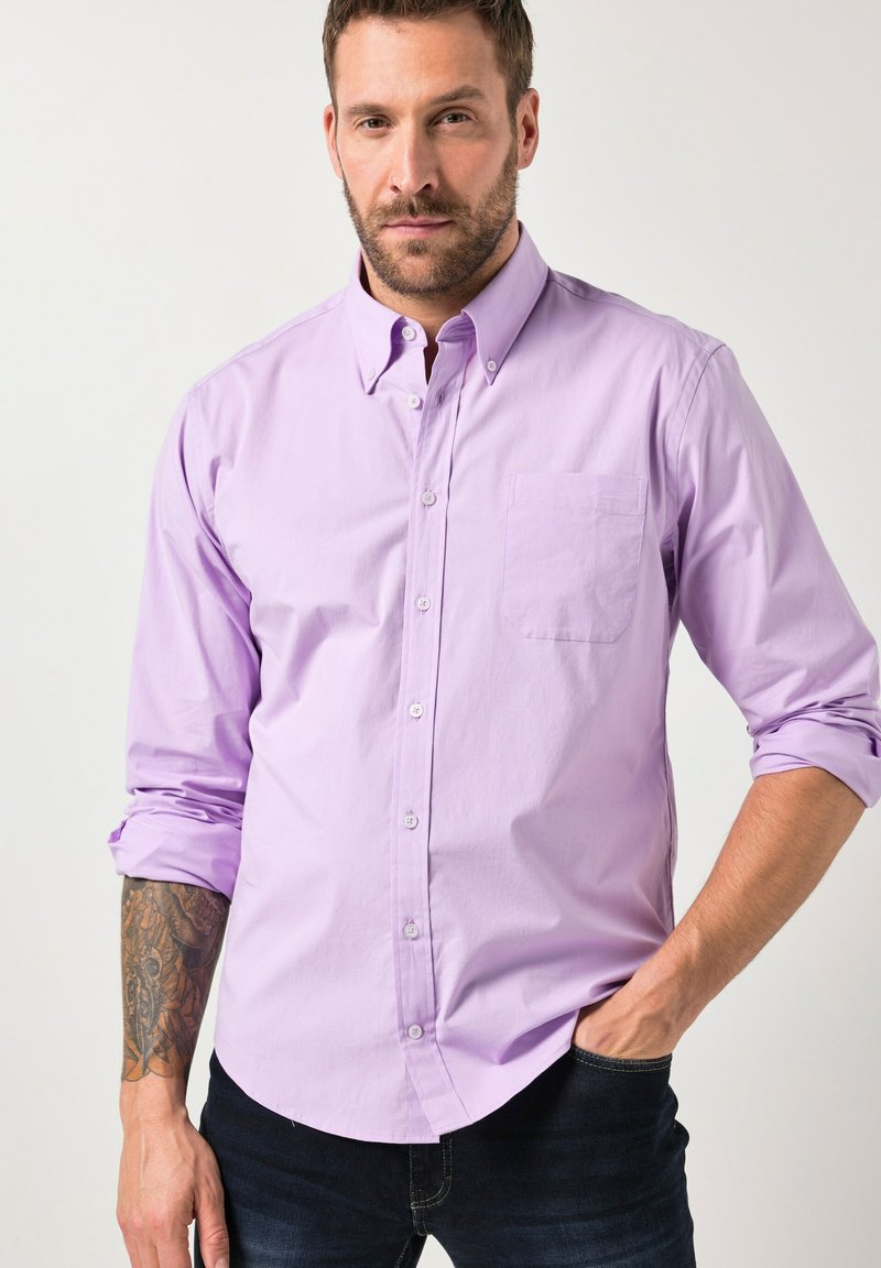 Mann mit Bart, der ein lavendelfarbenes Hemd mit hochgekrempelten Ärmeln und dunkle Jeans trägt, die Hand in der Tasche hat, mit einem sichtbaren Tattoo am linken Unterarm.