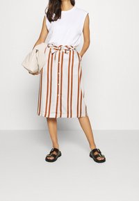 Haut sans manches blanc avec coutures d'épaule associé à une jupe beige arrivant aux genoux, ornée de rayures verticales orange, de boutons et d'une ceinture nouée à la taille.