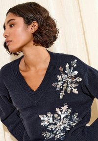 Pulover bleumarin cu decolteu în V. Prezintă decorațiuni cu fulgi de zăpadă din paiete pe partea stângă, creând un contrast texturat.