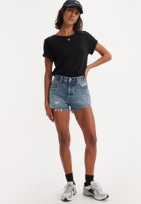 Camiseta negra de manga corta, pantalones cortos de mezclilla desgastados de cintura alta y zapatillas deportivas grises con calcetines negros. Estilo casual, ajuste relajado.