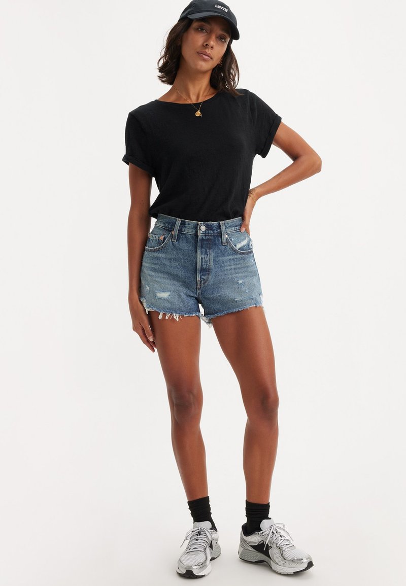 Camiseta negra de manga corta, pantalones cortos de mezclilla desgastados de cintura alta y zapatillas deportivas grises con calcetines negros. Estilo casual, ajuste relajado.