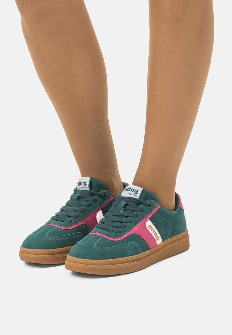 Teal suède sneakers met roze accenten en witte branding. Voorzien van een rubberen zool en donkere veters, met een lage ontwerp.