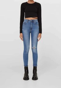 Top negro de manga larga y corte corto, jeans azul claro de tiro alto con detalles desgastados, y botines negros con suela gruesa.