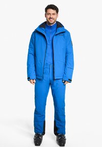 Giacca e pantaloni da sci blu realizzati in materiale impermeabile, con chiusura a zip frontale, cappuccio e dettagli di ventilazione. Gli stivali da sci completano l'outfit.