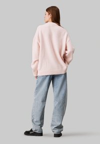 Maglione oversize rosa chiaro realizzato in materiale morbido e peloso; abbinato a jeans blu larghi e scarpe nere resistenti. Dettagli a coste semplici.