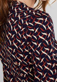 Marinblått tyg med ett snurrande mönster i röd och kräm. Plagget har en mjuk textur med en åtsittande design och rund hals.