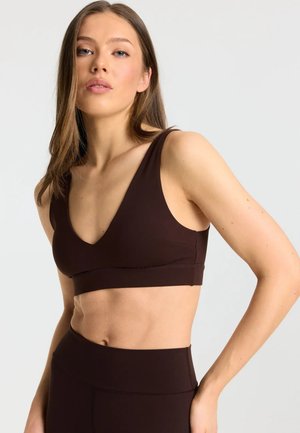 Femme aux longs cheveux bruns portant un soutien-gorge de sport marron foncé et des leggings taille haute sur un fond clair uni.