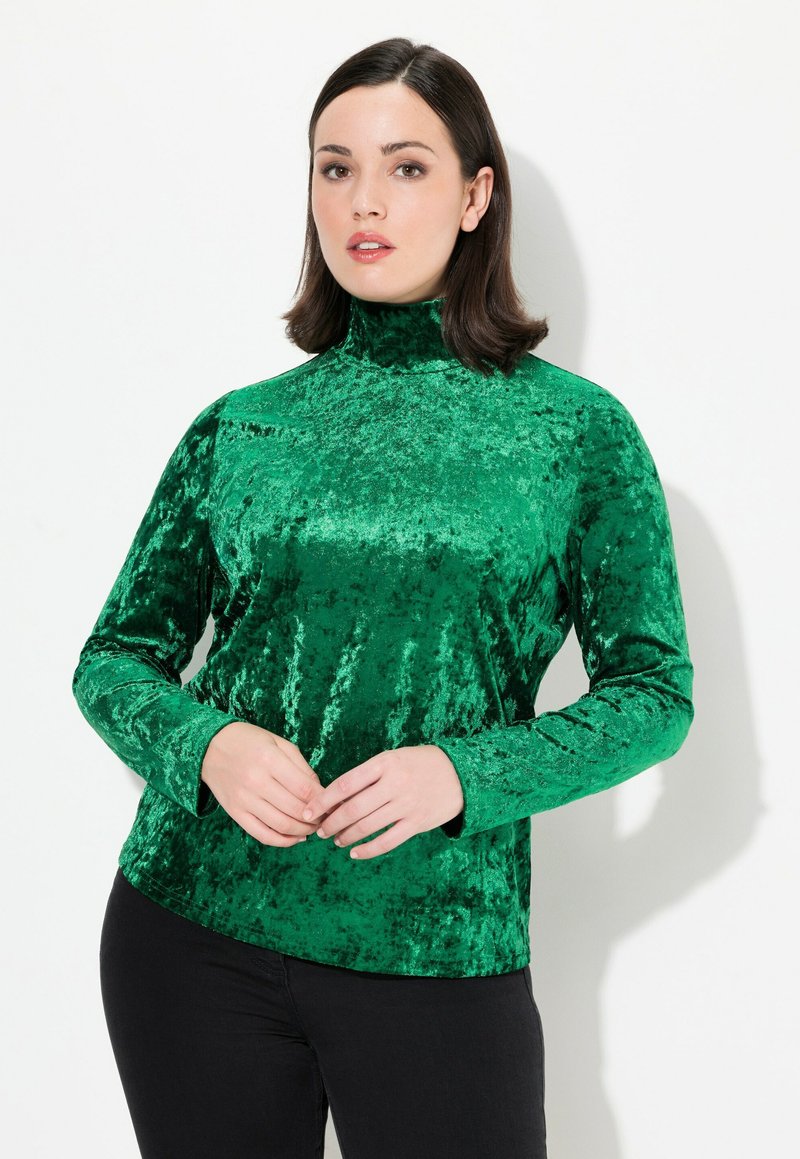 Dolcevita in velluto verde smeraldo con maniche lunghe, texture morbida, design aderente e leggera lucentezza, abbinato a pantaloni neri.