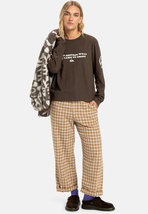Brown long-sleeve shirt with white text, plaid pants in orange and white, brown slippers, and a patterned gray blanket draped over one arm.
