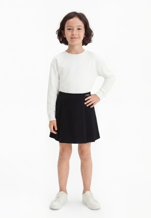 Jeune fille aux cheveux mi-longs portant une chemise blanche à manches longues, une jupe plissée noire et des baskets blanches, debout avec une main sur la hanche.