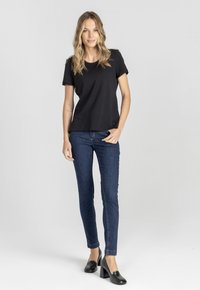Kobieta z falistymi blond włosami, ubrana w czarną bluzkę z krótkim rękawem, ciemnoniebieskie dopasowane dżinsy i czarne buty na słupku, stojąca na jednolitym tle.