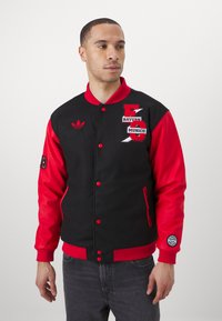 Giubbotto varsity nero e rosso con loghi ricamati, bottoni a pressione, polsini a costine e tasche laterali. Maniche lisce con un corpo testurizzato.