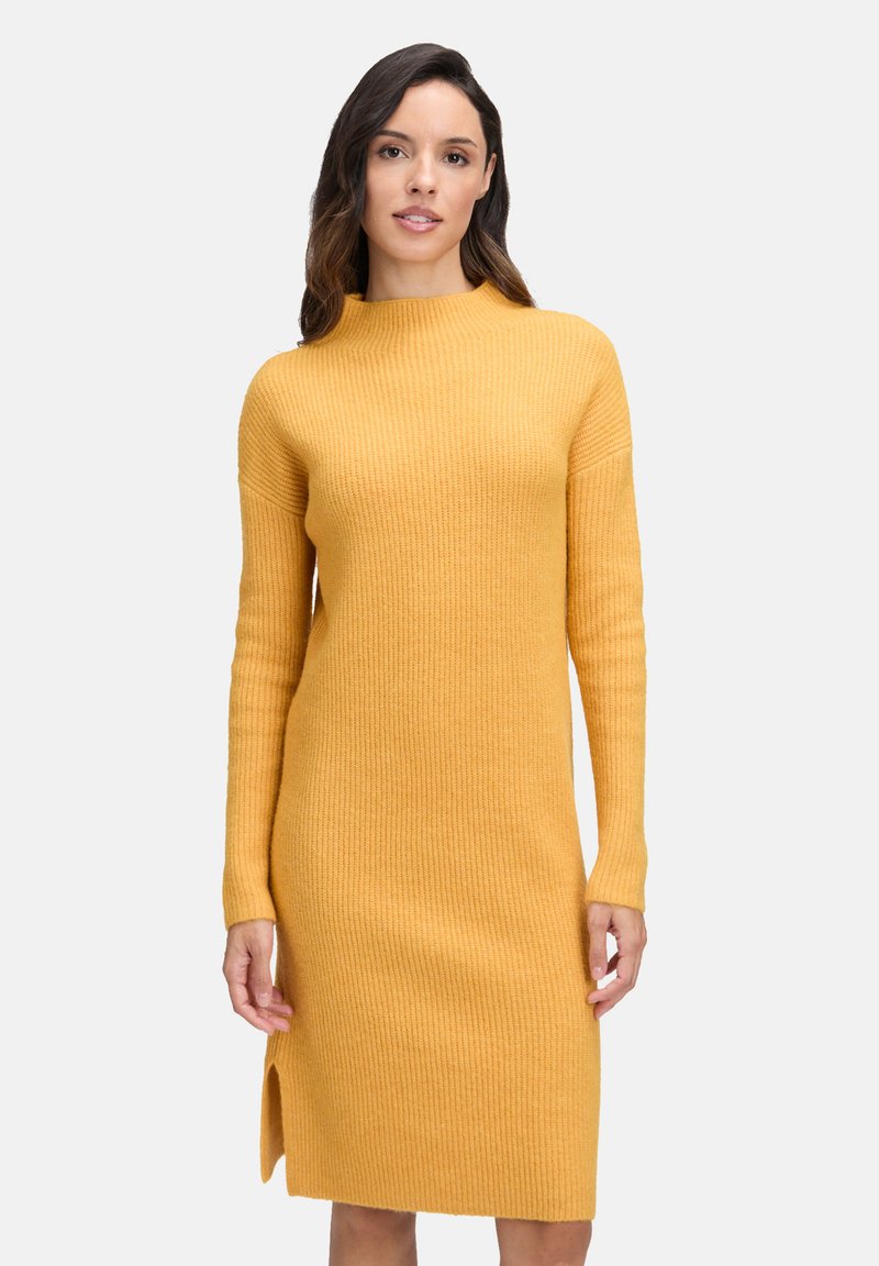 Eine Frau mit dunklen Haaren trägt ein senfgelbes, geripptes Langarmkleid mit einem seitlichen Schlitz und steht vor einem hellen, einfarbigen Hintergrund.