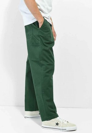 Person iført grønne cargobukser med flere lommer og hvide sneakers med sort stjernedetalje, stående med hænderne i lommerne.