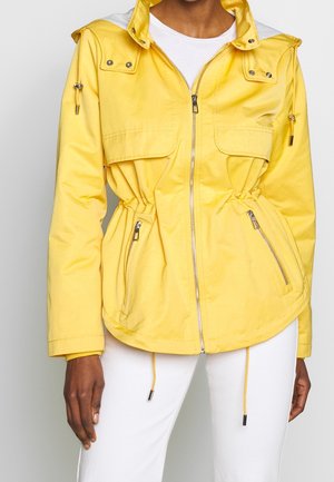 Veste jaune zippée avec poches avant, cordons de serrage réglables à la taille et col à boutons-pression, portée par-dessus une chemise blanche et un pantalon blanc.