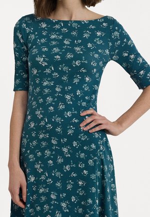 Frau trägt ein türkisfarbenes Kleid mit weißen Blumenmustern und ellbogenlangen Ärmeln, eine Hand auf der Hüfte, die andere locker an der Seite.