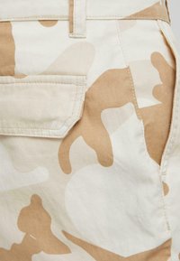 Beige camouflage shorts feature a back pocket with a flap, made from durable fabric with a smooth texture and subtle shine.