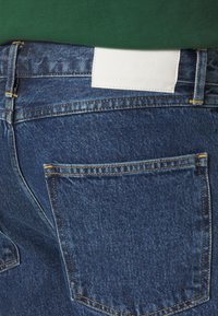 Vue arrière d'un jean denim bleu avec des coutures apparentes sur les poches et une étiquette rectangulaire blanche à la taille, porté avec un haut vert.