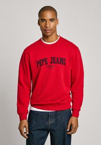 Sudadera roja de algodón con cuello redondo, que presenta el logo de "Pepe Jeans London" en azul marino. Puños y bajo de canalé, llevada sobre una camiseta blanca.