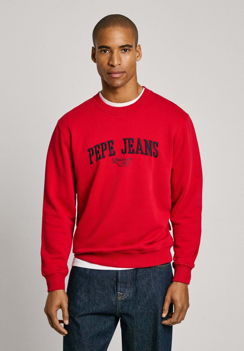 Sudadera roja de algodón con cuello redondo, que presenta el logo de "Pepe Jeans London" en azul marino. Puños y bajo de canalé, llevada sobre una camiseta blanca.