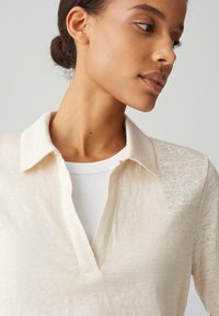 Chemise col bénitier beige clair en tissu texturé, avec un design à enfiler et un col en V, superposée sur un haut blanc à côtes.
