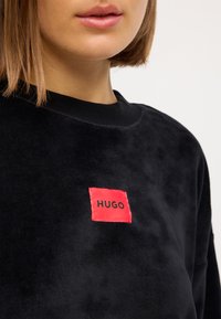 Crna flis dukserica koja ima kvadratnu crvenu oznaku s natpisom "HUGO" u crnom slovu na prsima, okrugli izrez i mekanu teksturu.