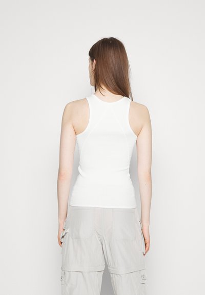 Hvid tætsiddende tanktop med rund halsudskæring og racerback-design, der har en ribbet tekstur og glat stof, båret med lysegrå cargo-bukser.