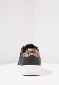 Baskets noires à texture lisse avec une semelle blanche, présentant un accent en tissu à motif coloré sur le talon. Design globalement minimaliste.