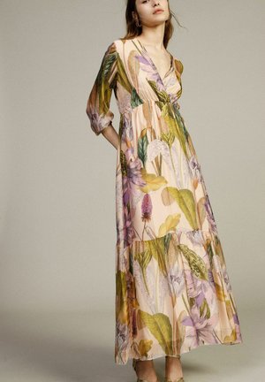 Donna che indossa un lungo vestito floreale fluente con foglie e fiori verdi, viola e gialli, in piedi davanti a uno sfondo beige semplice.