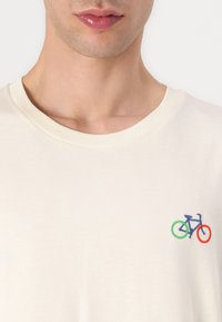 Krém színű pamut póló, apró hímzett kerékpár mintával kék, zöld és piros színekben. Kerek nyakú és rövid ujjú.