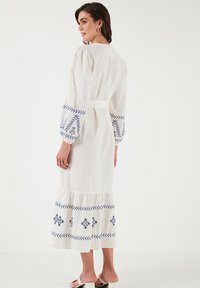 White embroidered dress featuring blue patterns, long puff sleeves, a fitted waist belt, and a ruffled hem. Casual and breathable fabric.