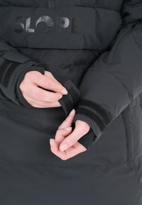 Svart puffjacka med SLOPE-logotyp. Har justerbara kardborrehandledningar och ett slätt, texturerat tyg med sömmar och isolering.