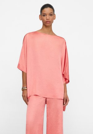 Vrouw draagt een loszittende, koraalroze satijnen top en bijpassende wijde broekspijpen, met gouden armbanden en haar naar achteren gebonden tegen een effen witte achtergrond.