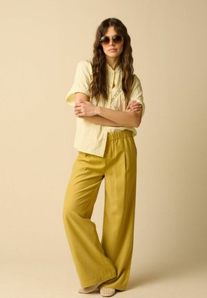 Femme aux longs cheveux ondulés portant de grandes lunettes de soleil rondes, une blouse crème à manches courtes, un pantalon large couleur moutarde et des chaussures tissées beige, debout, les bras croisés.