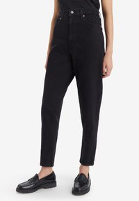 Person som bär högmidjade svarta jeans och svarta läder loafers, står med avslappnade händer vid sidorna mot en vit bakgrund.