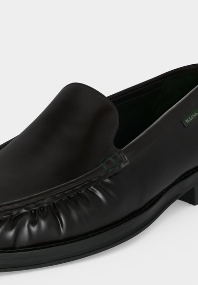 Mocassin en cuir noir à enfiler, bout arrondi, détails froncés sur les côtés et petit talon sur fond blanc uni.