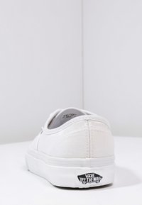 White canvas sneaker with a low-top design, rubber sole, and black logo tag on the heel. Features stitching accents and plain texture.