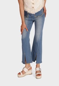 Jeans a zampa d'elefante in denim blu chiaro con applicazioni floreali dorate sulle tasche, caratterizzati da spacchi laterali all'orlo, abbinati a sandali con zeppa bianchi.