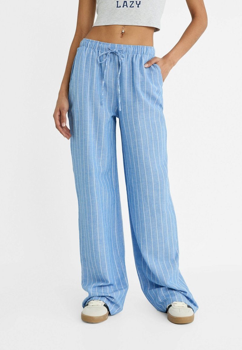 Femme portant un pantalon bleu clair à jambes larges avec des rayures verticales blanches et une taille avec cordon, associé à des baskets blanches et un top court gris.