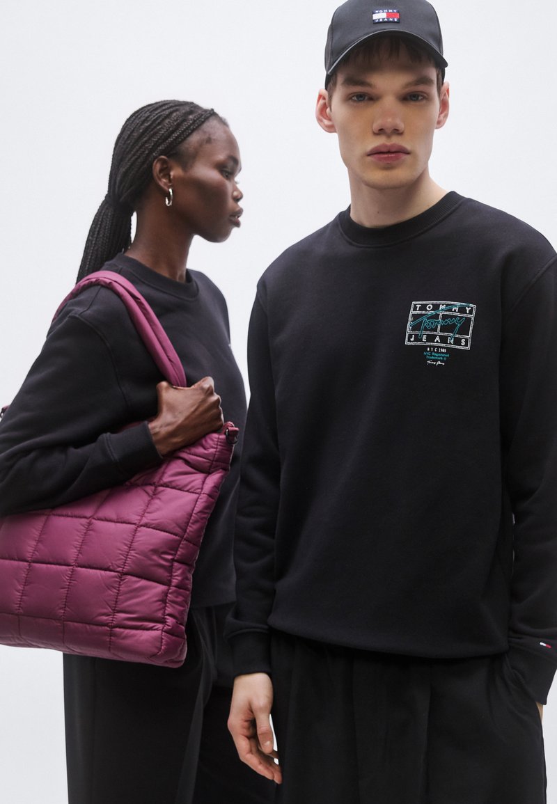 Black sweatshirts with printed logos, a quilted purple bag, and a black cap. Braided hair visible, with hands holding the bag.