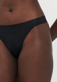 Zwarte bikini onderkant met een gladde, zachte textuur, voorzien van een scalloped randontwerp. Minimale bedekking, klassieke low-rise vorm.