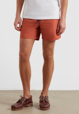 Man wearing rust-colored shorts, white t-shirt, and brown leather boat shoes standing on a concrete floor against a white wall.